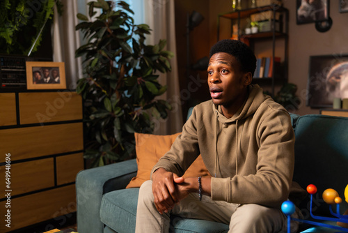 Fearful and anxious expression of an African man in his living room, gripping his remote control tightly and showing clear signs of distress