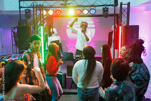 Clubbers crowd dancing at techno music concert and partying in nightclub. Dj singing and live performing on stage while diverse people moving and having fun on club dancefloor