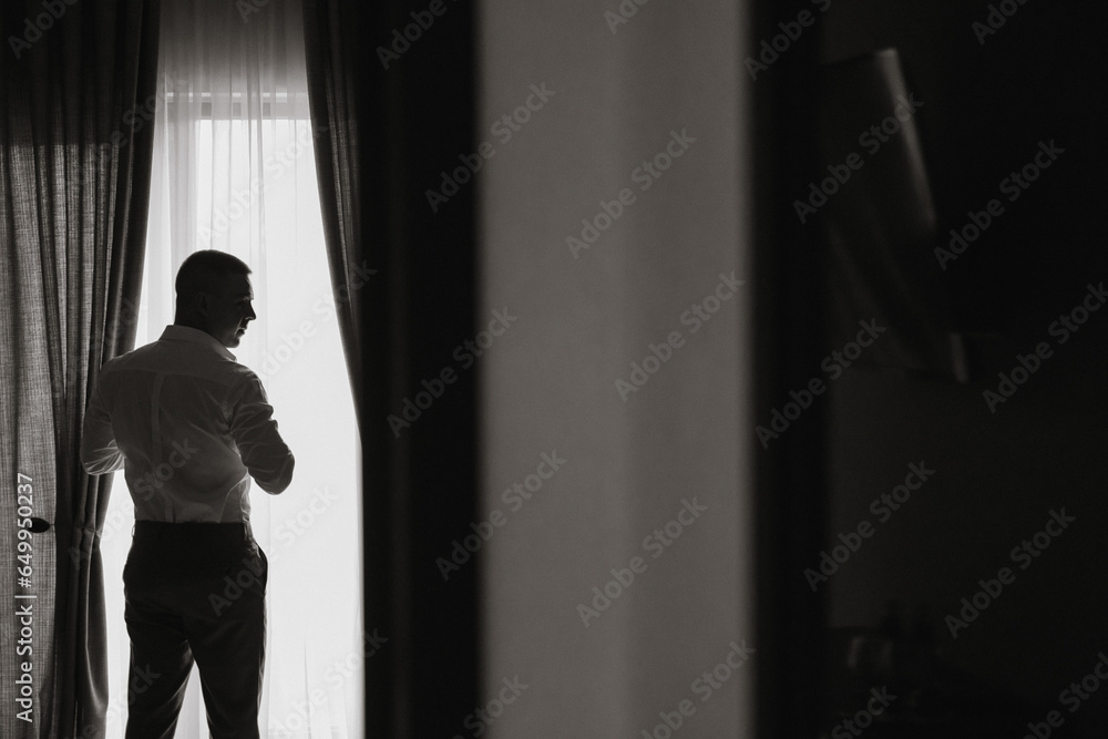 Relaxed man puts on white shirt in hotel room with natural light. The groom wears a shirt and prepares for the wedding. Free space
