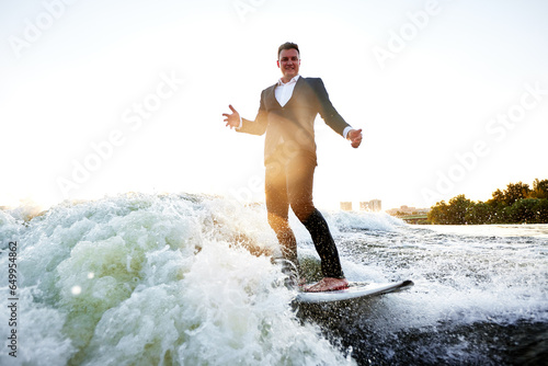 Young man in classic suit rides a wakeboard on the river or lake near city. Clerk escaped from a stuffy office to take up his favorite active sport. Best summer leisure after routine work. Sun flare.