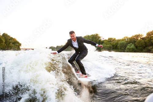 Young man in classic suit rides a wakeboard on the river or lake near city. A careless clerk escaped from a stuffy office to take up his favorite active sport. Best summer leisure after routine work.