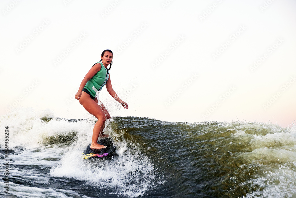 Young beautiful sporty girl in a green life jacket rides a wakeboard on a river or lake near city. Happy athlete girl glides on the wave like a real surfer. Active lifestyle, healthy hobby.