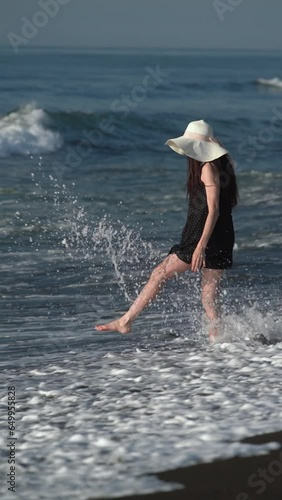 Playful barefoot woman with long legs standing on beach, kicking and raising splashes in waves crashing seashore. Unrecognizable person in black polka dot dress, straw hat during summer beach holidays