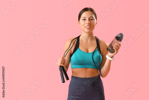 Sporty young woman with jumping rope and water on pink background