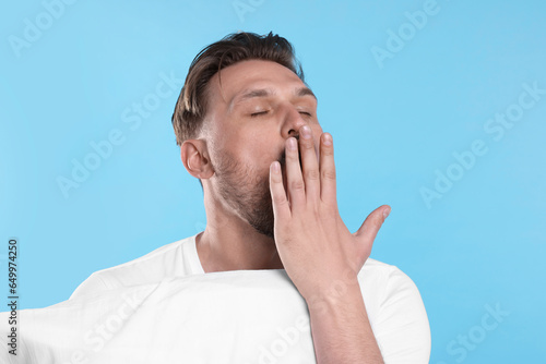 Sleepy man with pillow yawning on light blue background. Insomnia problem