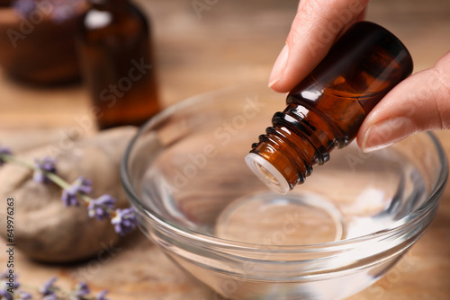 Woman dripping lavender essential oil from bottle into bowl at wooden table, closeup. Space for text
