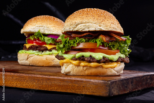 2 giant hamburgers with an iron basket containing fries, on top of a wooden board and all on a table with black fabric and a black background