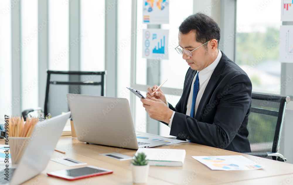 Asian professional successful male businessman manager entrepreneur in formal suit and glasses sitting at working laptop computer at desk in company office Asian professional successful male businessman manager entrepreneur in formal suit and glasses sitting at working laptop computer at desk in company office