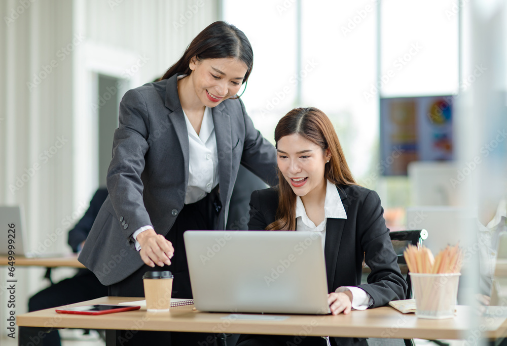 Asian professional successful female businesswoman supervisor mentor in formal suit standing smiling pointing help advising new colleague working with paperwork document at working station in office Asian professional successful female businesswoman supervisor mentor in formal suit standing smiling pointing help advising new colleague working with paperwork document at working station in office