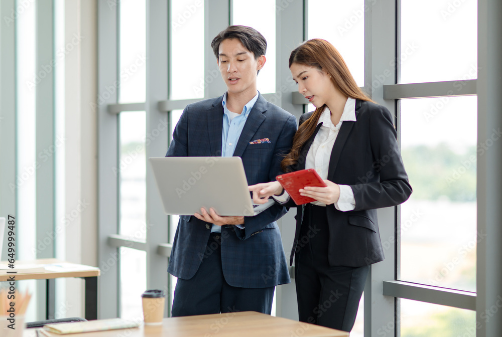 Asian professional successful female businesswoman and male businessman colleague employee in formal suit standing smiling together browsing surfing internet online via laptop and tablet in office