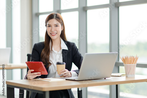 Portrait shot of Asian professional successful female businesswoman secretary employee in formal suit sitting smiling look at camera taking break drink coffee browsing tablet online in company office