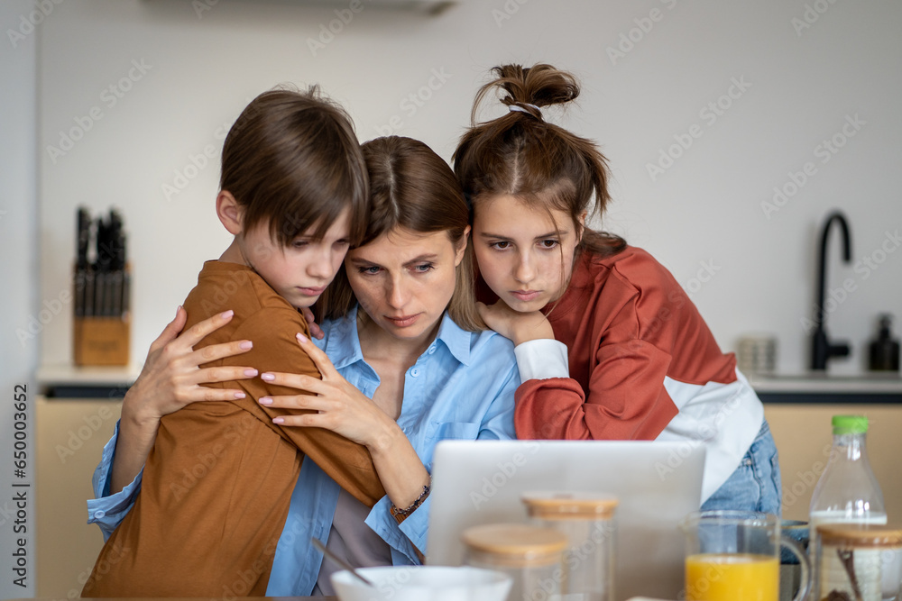 Teenagers children support mother in kitchen looking at laptop screen ...