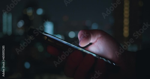 Close up person use mobile phone at night time on urban light background. Cinematic film look of male hand use mobile phone, tapping and scrolling news feed on app on his phone at night