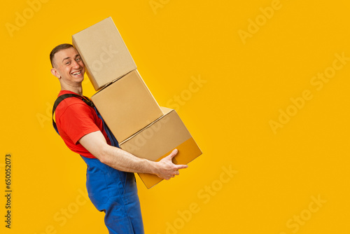 Laughing loader carries large boxes. Yellow background. Portrait of young man with pyramid of cardboard boxes in his hands. Copy space, mock up.