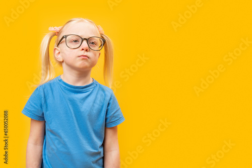 Portrait of sad little blonde girl in glasses and looks seriously. Preschooler on yellow background. Copy space © somemeans