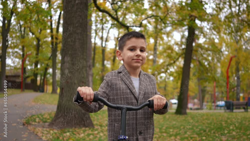 Smiling confident boy looking at camera strolling with scooter on park alley. Portrait of positive Caucasian kid posing in slow motion walking outdoors on overcast autumn day