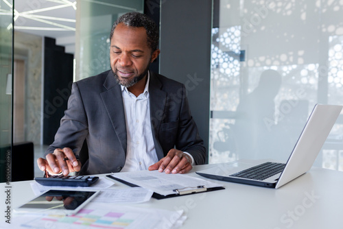 Successful satisfied financier behind paper work, afro american senior experienced filling out documents, using calculator for calculation, businessman behind paper work inside office smiling. photo