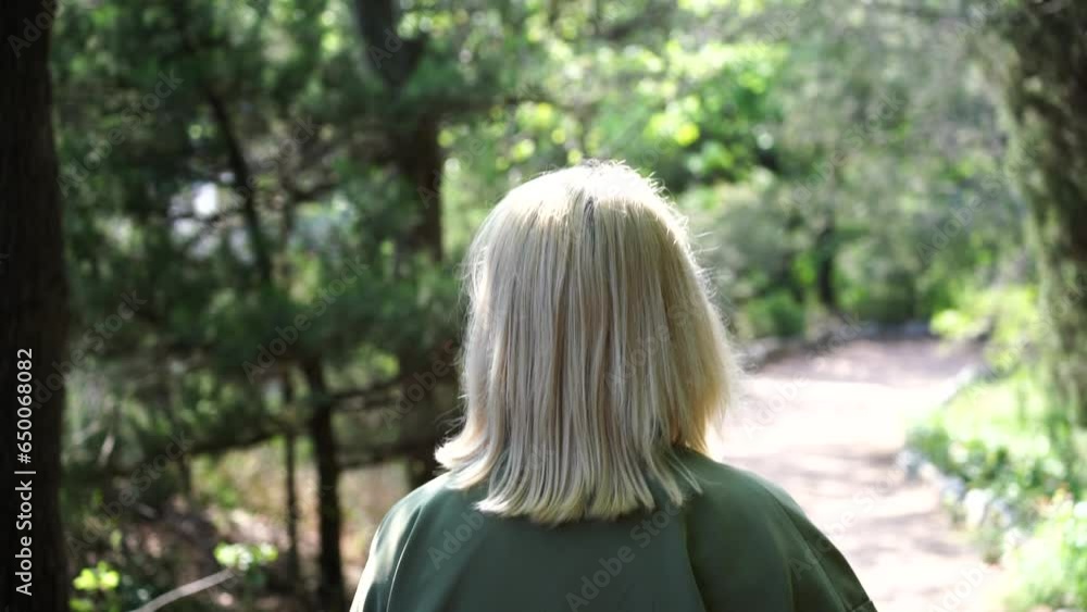Blonde walking in the park. Happy young woman wearing. Happy young woman in a green jacket walks forward in the park, rear view, slow motion Blonde walking in the park. Happy young woman wearing. Happy young woman in a green jacket walks forward in the park, rear view, slow motion