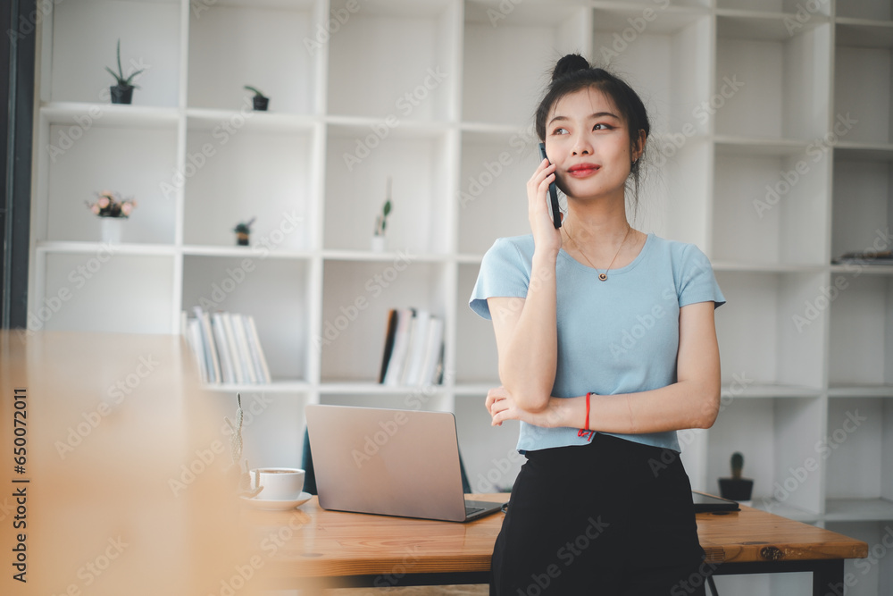 Attractive young woman talking on the mobile phone and smiling while standing at her working place in office and looking at laptop. Attractive young woman talking on the mobile phone and smiling while standing at her working place in office and looking at laptop.