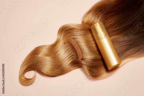 Ply of shiny, wavy, blonde hair with wooden brush and hair product, conditioner, mask over light background. Concept of hair care, organic products, natural beauty, cosmetics. Ad. Poster.