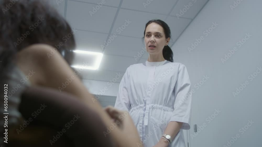 Female doctor stands in clinic corridor, consults patient in wheelchair. Healthcare specialist talks to woman with physical disability about medical examination. Hallway of modern medical facility. Female doctor stands in clinic corridor, consults patient in wheelchair. Healthcare specialist talks to woman with physical disability about medical examination. Hallway of modern medical facility.
