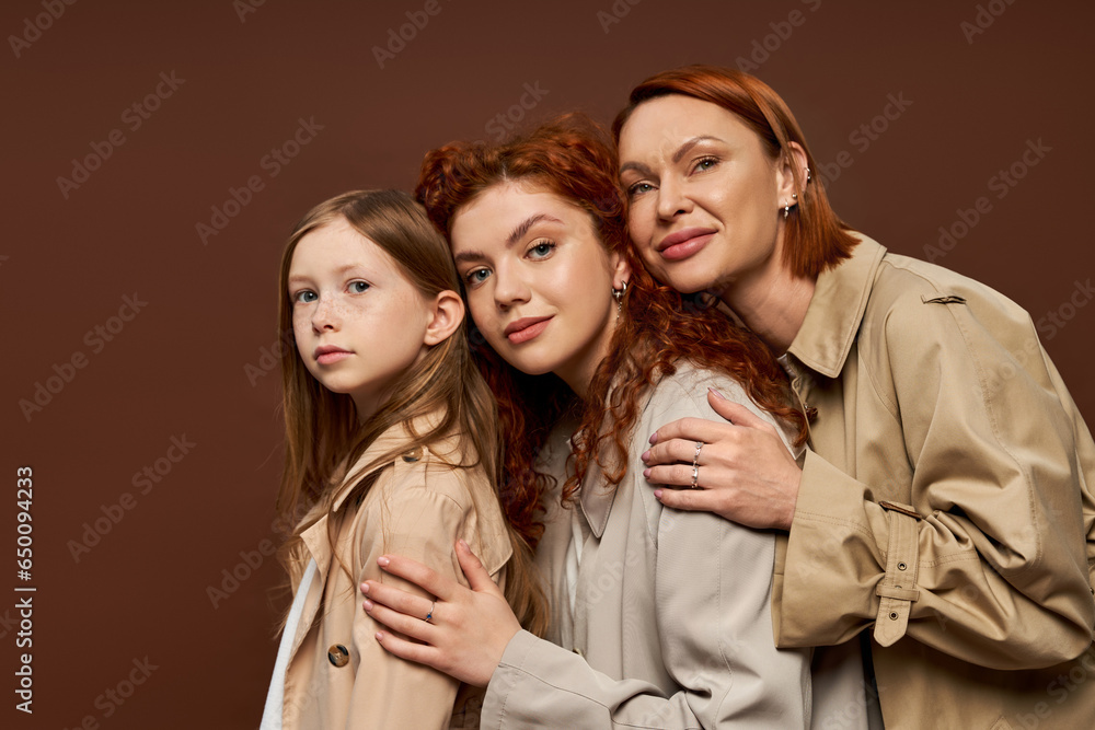family of three female generations with red hair posing in beige coats, hugging on brown background family of three female generations with red hair posing in beige coats, hugging on brown background