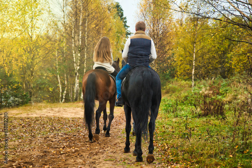 Rear view of couple man and woman on horses walk in fall forest, rural scene. From behind riders on horseback in autumn park, countryside. Leisure activity and recreation concept. Copy ad text space