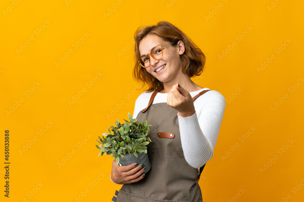 Young Georgian woman holding a plant isolated on yellow background making money gesture Young Georgian woman holding a plant isolated on yellow background making money gesture