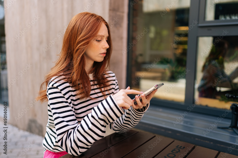 Φωτογραφία Side view of pensive young woman looking at phone online dating swiping left and right on a dating app, liking some of profiles and not liking some, sitting at table in outdoor cafe