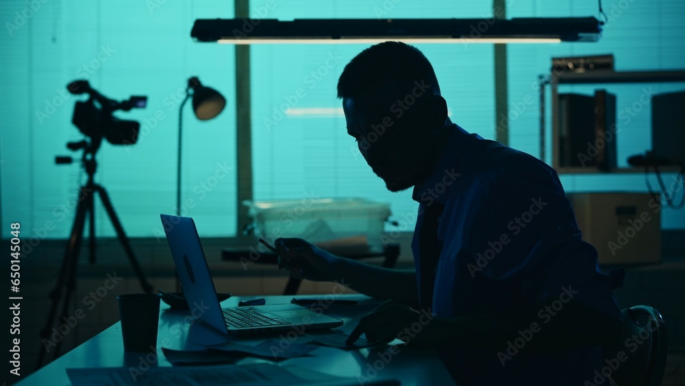 Αυτοκόλλητο Medium silhouette shot of a detective, policeman sitting in the interrogation room, smoking, working on a laptop and studying the evidence