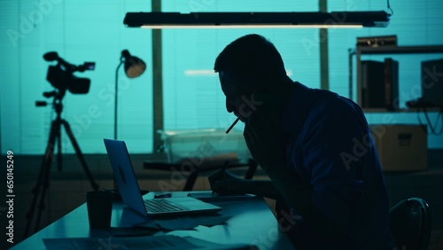 Medium silhouette shot of a detective, policeman sitting in the interrogation room, smoking, working on a laptop and studying the evidence. photo