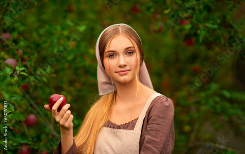 Portrait of beautiful blonde girl in medieval dress and apron taking red apple. Young worker standing in garden.Warm colorful art work.
