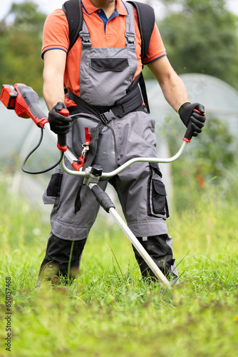 Man in protective clothes mowing green grass with cordless lawn trimmer in garden