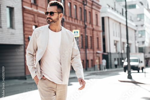 Portrait of handsome confident stylish hipster lambersexual model. Sexy modern man dressed in elegant suit. Fashion male posing in the street background in Europe city at sunset. In sunglasses