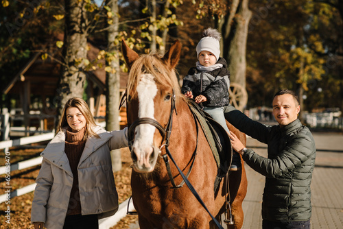 Kid riding a horse. Family having fun spring vacation on farm ranch. Little boy ride in saddle on horse. Happy father, mother, son caress horse outdoors. Dad, mom, child cuddling animal pet at sunset. © Serhii