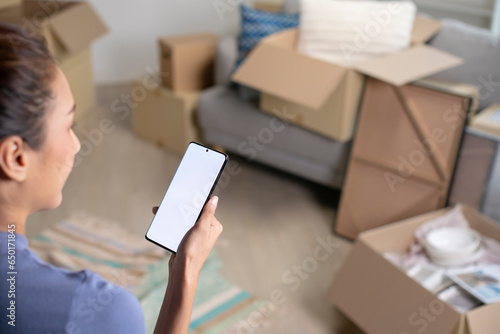 Young Asian woman sitting in new house using smartphone buying decorations online after moving in.