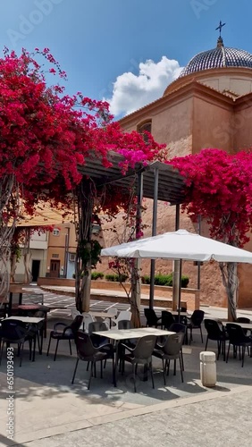 The center of the Spanish city of Benicasim in spring. Red flowers against a background of blue sky and church. The historic city center by the Mediterranean Sea, popular in Spain.