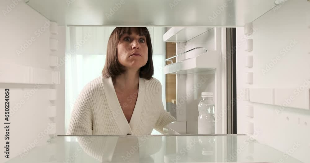 A woman opens her fridge, only to find it disappointingly empty. She retrieves a water bottle, face reflecting slight dismay at the lack of available food. A woman opens her fridge, only to find it disappointingly empty. She retrieves a water bottle, face reflecting slight dismay at the lack of available food.