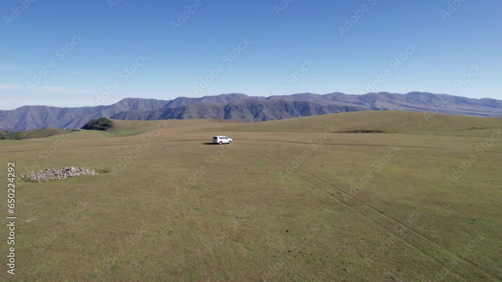 A white SUV is driving through mountain green fields. In the distance there are high peaks with snowy peaks. Dirty road. Blue sky. A dog runs and shepherd rides. Mountainous terrain, sometimes forest A white SUV is driving through mountain green fields. In the distance there are high peaks with snowy peaks. Dirty road. Blue sky. A dog runs and shepherd rides. Mountainous terrain, sometimes forest