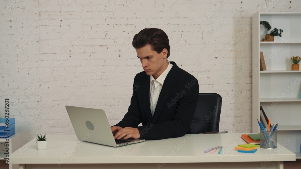 Medium shot of a young businessman sitting at the table in an office, working on his laptop, focused, consentrated and seious. Medium shot of a young businessman sitting at the table in an office, working on his laptop, focused, consentrated and seious.