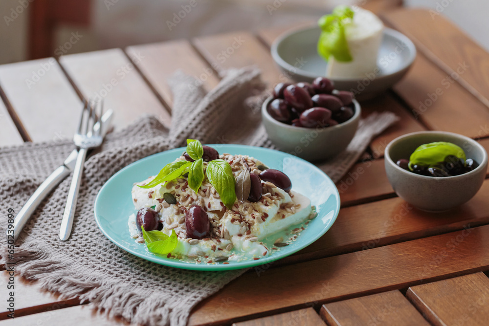 Feta cheese sliced on blue plate with sesame, flax seeds, served with black olives and basil leaves. Delicious and healthy breakfast on terrace