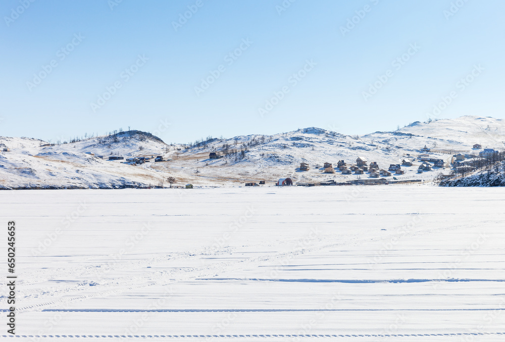 Baikal Lake in winter. View from the snow-covered ice of the Kurkut Bay to tourist wooden houses on the shore on a sunny February day. Winter travel on lake ice concept. Natural cold background - obrazy, fototapety, plakaty 