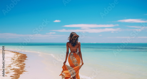 A young and beautiful african american woman is walking on the sand next to the waterline with a dress on a tropical beach with a calm ocean - summer weather beach walk relaxing photo