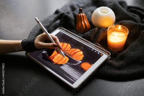 Girl's hand in special glove draws still life picture with pumkins on electronic tablet near burning candle. The concept of inspiration, creativity, modern art. Halloween, Thanksgiving day
