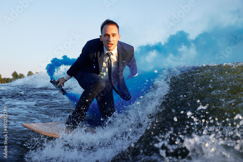 Canvas Print Young man in classic suit with blue smoke bomb in his hand rides wakeboard on a lake near city