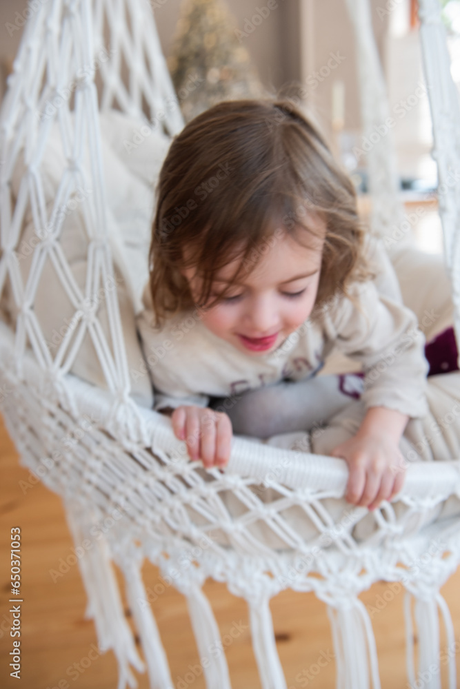 Fototapeta premium Defocused close-up portrait of funny little girl riding on swing on macrame chair at home.