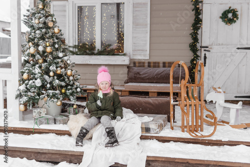 A little girl in winter clothes is sitting on the porch of a house decorated for the Christmas holidays. Christmas holidays for a child.