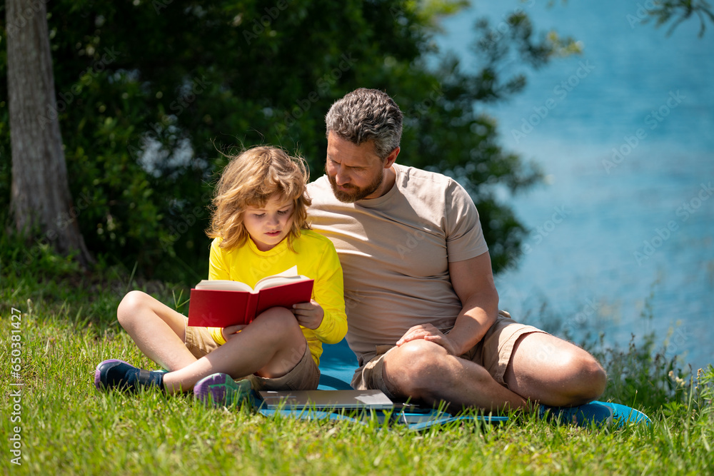 Father reading book with son in park, preparing school homework together, parenting. Summer lifestyle. Parenting and childhood concept. Little boy learning with father in outdoors garden.