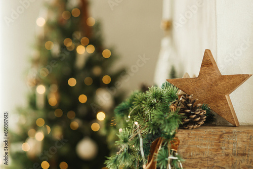 Stylish christmas wooden star, spruce branches on fireplace mantel close up on background of christmas tree lights. Festive rustic fireplace decor in modern farmhouse living room photo