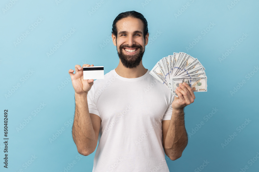 Man with beard wearing white T-shirt holding credit card and dollar banknotes, feeling rich with lot of money, happy to get bank loan for shopping. Indoor studio shot isolated on blue background. Man with beard wearing white T-shirt holding credit card and dollar banknotes, feeling rich with lot of money, happy to get bank loan for shopping. Indoor studio shot isolated on blue background.